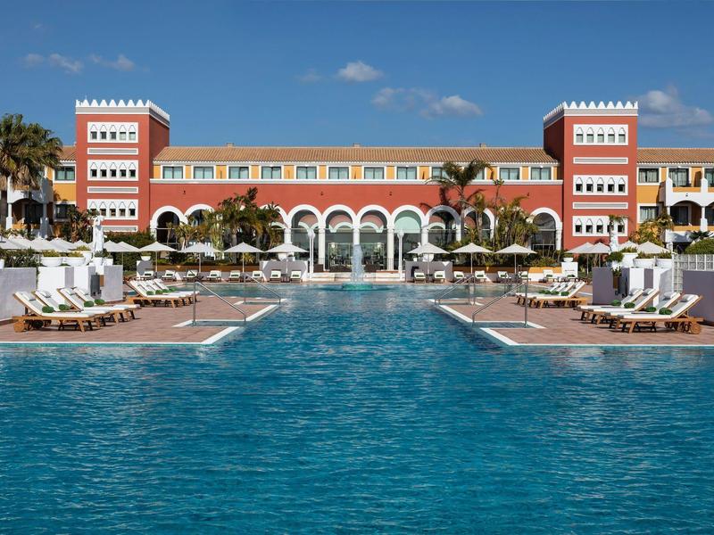 Großes Hotel mit rotem Backsteingebäude, weißem Pool und Liegestühlen unter blauem Himmel.