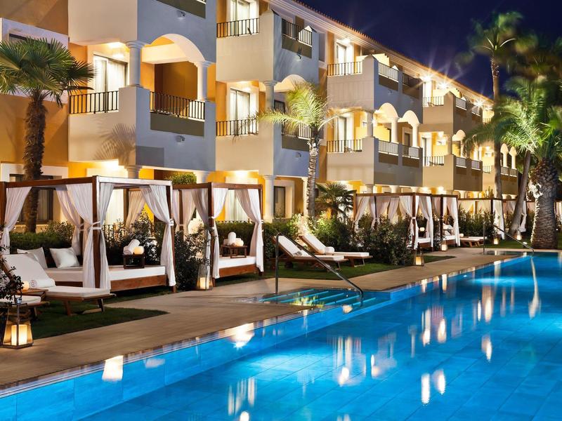 Beleuchteter Poolbereich mit Liegen und Pavillons vor einem mehrstöckigen Hotelgebäude bei Nacht.