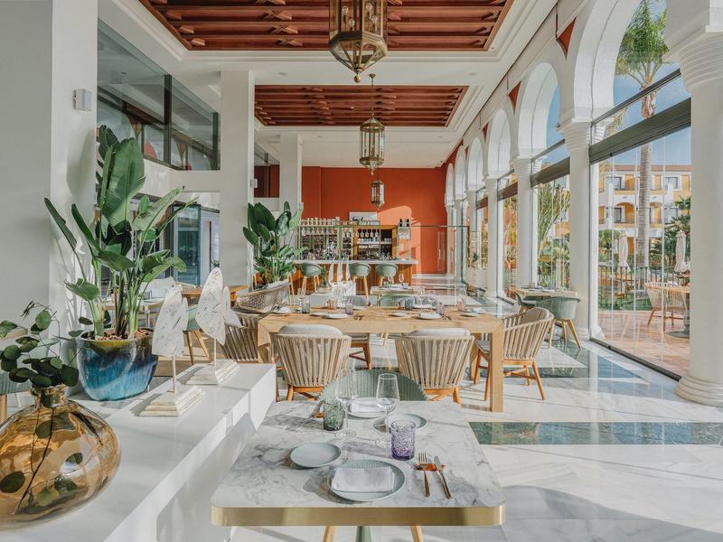 Helles Restaurant mit großen Fenstern, Holztischen, Rattan-Stühlen und grünen Pflanzen.