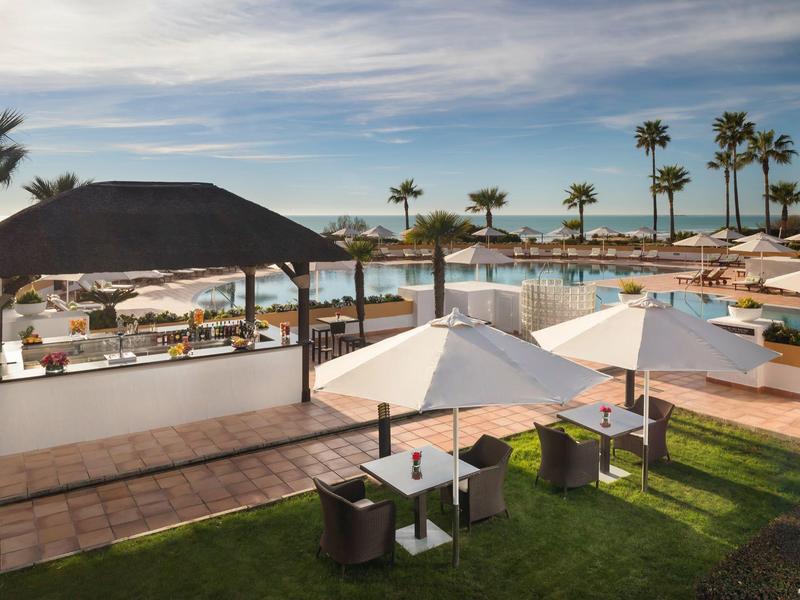 Hotelpool mit Sonnenschirmen, Liegen, Palmen und Blick auf das Meer bei bewölktem Himmel.