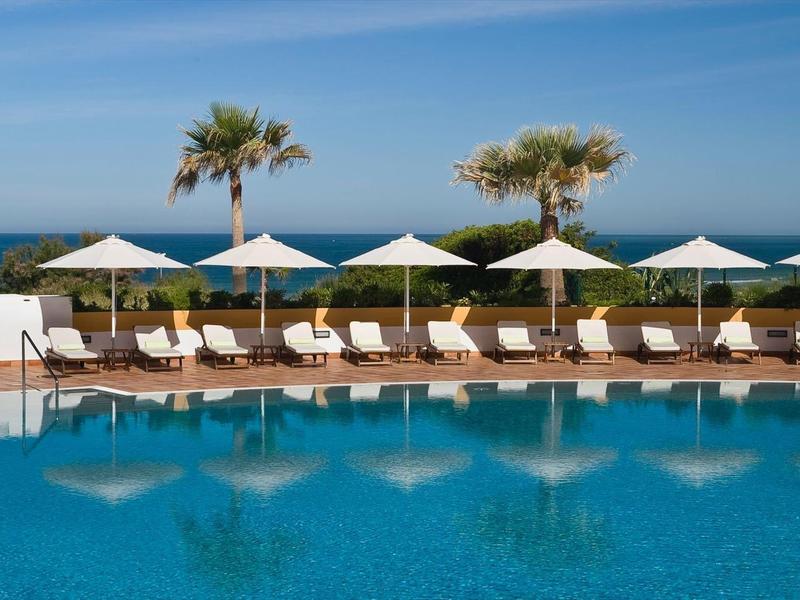 Großer blauer Pool mit weißen Liegestühlen, Sonnenschirmen und Palmen, Meer im Hintergrund.