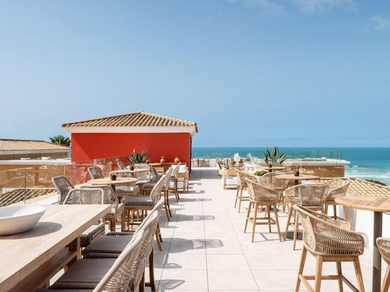 Terrassenrestaurant am Meer mit Holztischen, Stühlen und blauem Himmel.