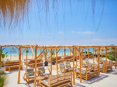 Strand mit Holzliegen unter Bambusdächern, weißer Sand, blaues Meer und klarer Himmel im Hintergrund.