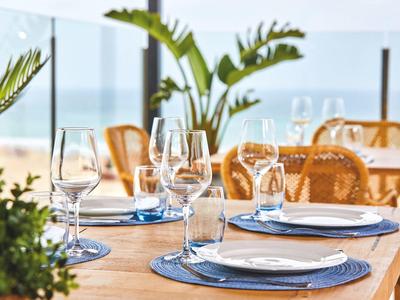 Ein gedeckter Holztisch mit Weingläsern, Teller und blauen Platzsets in hellem, offener Raum.