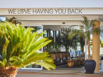Hotelterrasse mit Palmen und Stühlen unter einem Dach mit der Aufschrift "We love having you back".