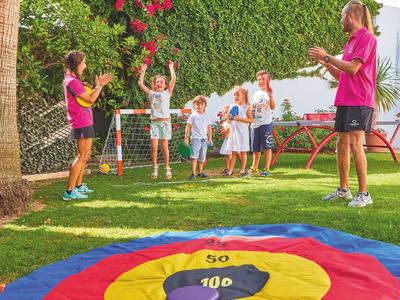 Kinder spielen Fußball im Garten mit zwei Erwachsenen, grüner Hecke und buntem Zielkreis auf Gras.