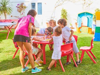 Kinder sitzen und stehen an bunten Tischen und Stühlen im Garten bei Sonnenschein und Spielen.