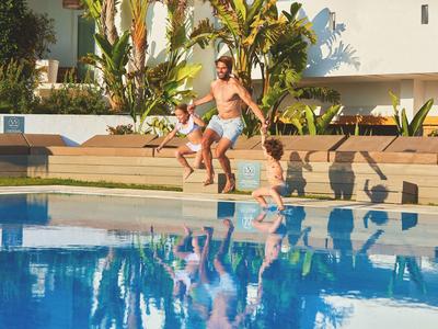 Drei Kinder springen vom Beckenrand in einen blauen Swimmingpool an einem sonnigen Tag.