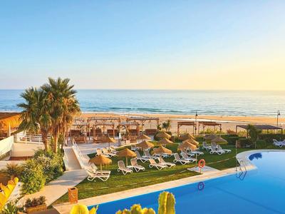 Hotelpool mit Liegestühlen, Palmen und Strand im Hintergrund bei Sonnenuntergang.