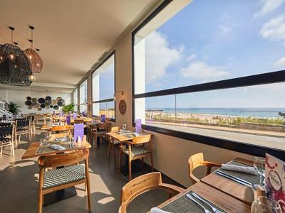 Restaurant mit Holztischen und Stühlen, großen Fenstern und Blick aufs Meer bei Tageslicht.