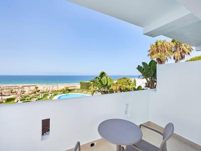 Balkon mit Tisch und Stühlen, Blick auf Meer, Pool und Pflanzen bei klarem Himmel.