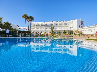 Großer blauer Pool vor weißem Hotelgebäude mit Palmen und klarem Himmel im Hintergrund.