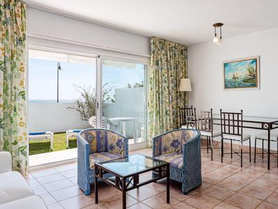Helles Wohnzimmer mit großen Fenstern, blauen Sesseln, Esstisch, Blumen und Blick auf Meer.