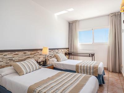 Hell eingerichtetes Zimmer mit zwei Einzelbetten, weißer Wand, Fenster und braunem Fliesenboden.
