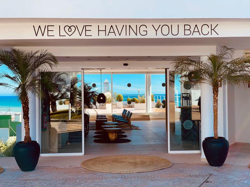 Eingang eines Resorts mit Glasfront, Palmen und Blick auf das Meer im Hintergrund.