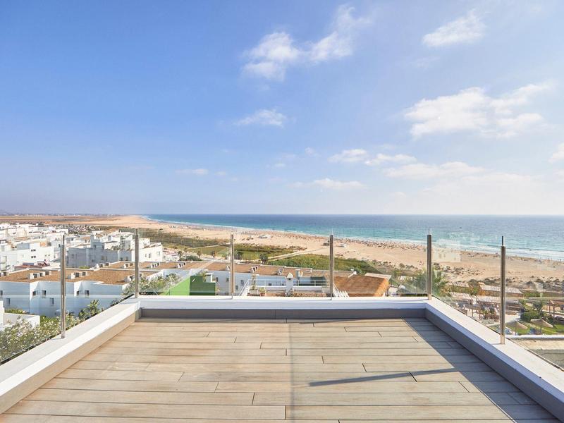 Blick von einer Terrasse mit Glasgeländer auf das Meer und einen Sandstrand bei klarem Himmel.