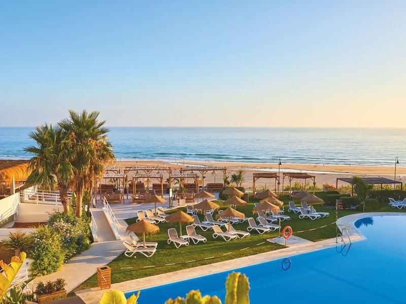 Hotelpool mit Liegestühlen neben Strand und Meer bei Sonnenuntergang.