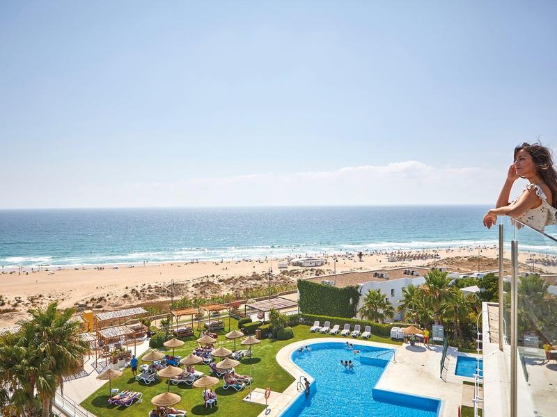 Blick auf einen Hotelpool, Garten mit Liegestühlen und einen Sandstrand am Meer.