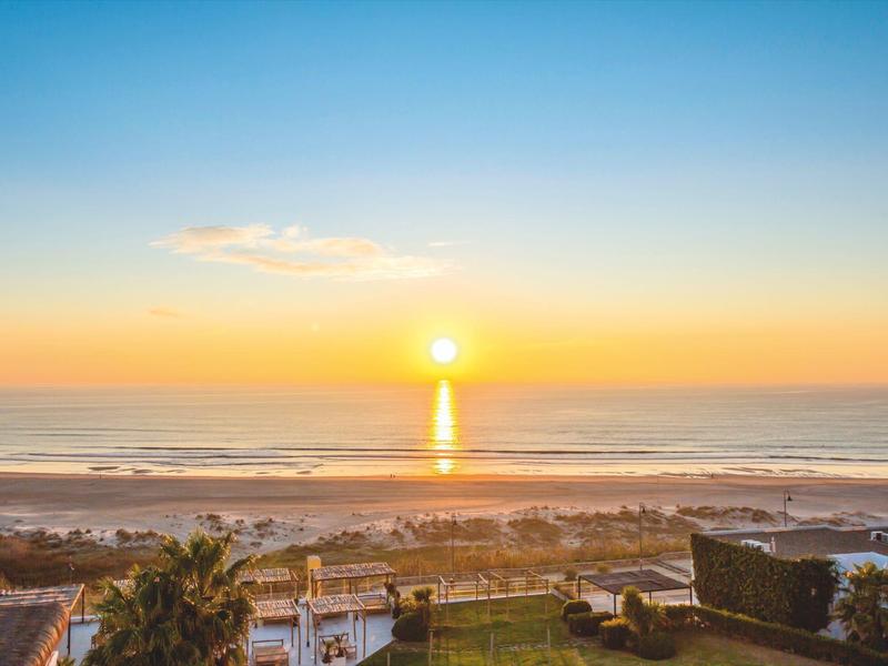Sonnenuntergang über dem Meer mit Strand, Wellen und Häusern im Vordergrund.