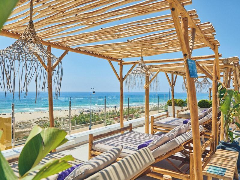 Lehnstühle unter einer Holzpergola mit Blick auf einen sonnigen Sandstrand und das blaue Meer.