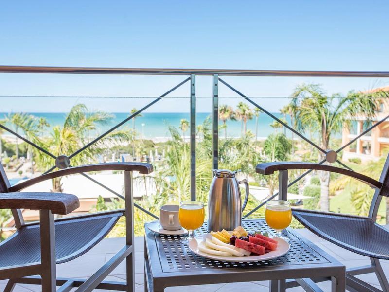 Balkon mit zwei Stühlen, Tisch mit Frühstück und Blick auf Palmen und Meer bei klarem Himmel.