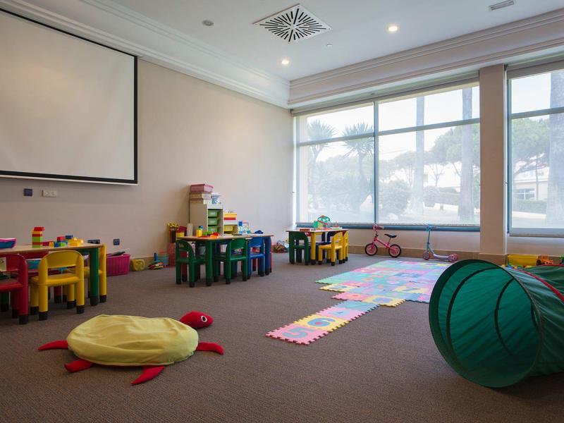 Ein heller Spielraum mit bunten Tischen, Spielzeug, Sitzkissen und großem Fenster.