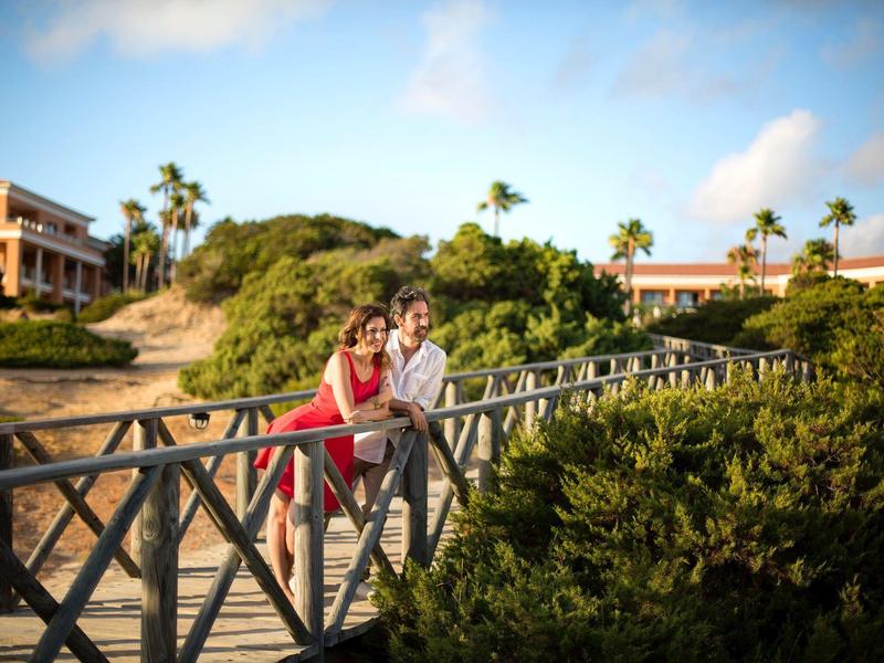 Paar lehnt auf Holzbrücke, umgeben von Grün, Häusern und blauem Himmel im Hintergrund.