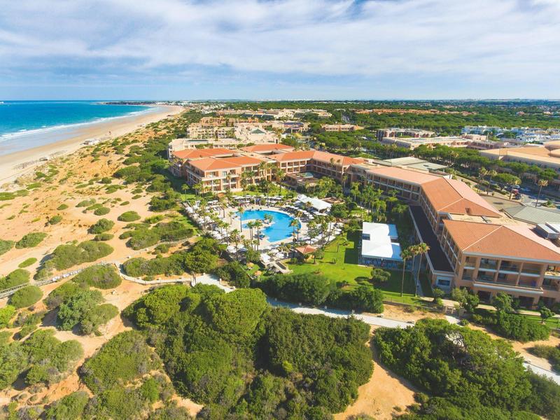 Große Küstenanlage mit Pool, umliegender Vegetation und weitem Sandstrand unter blauem Himmel.