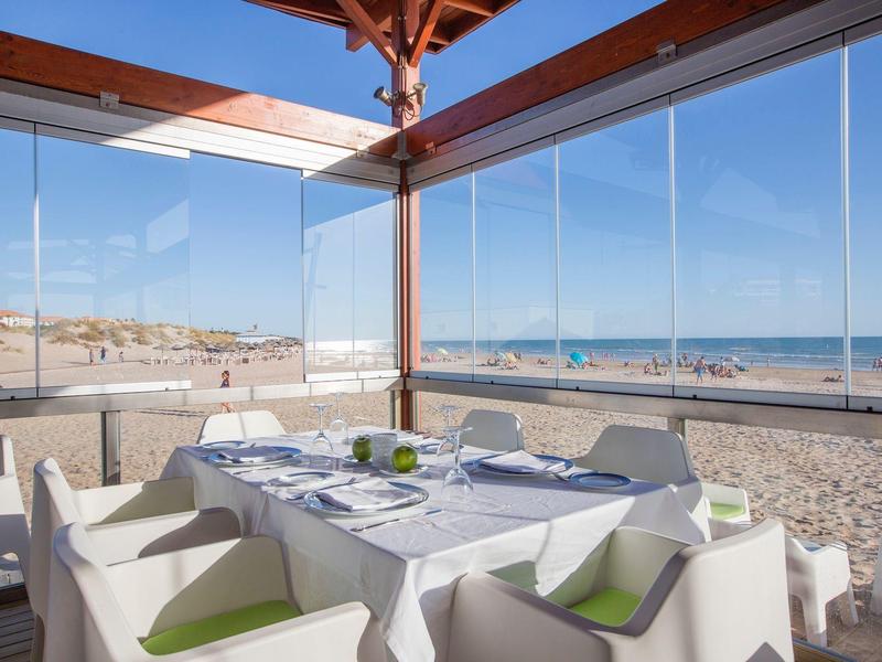 Strandrestaurant mit gedecktem Tisch und weißen Stühlen, Palmen und Meer im Hintergrund bei Sonne.