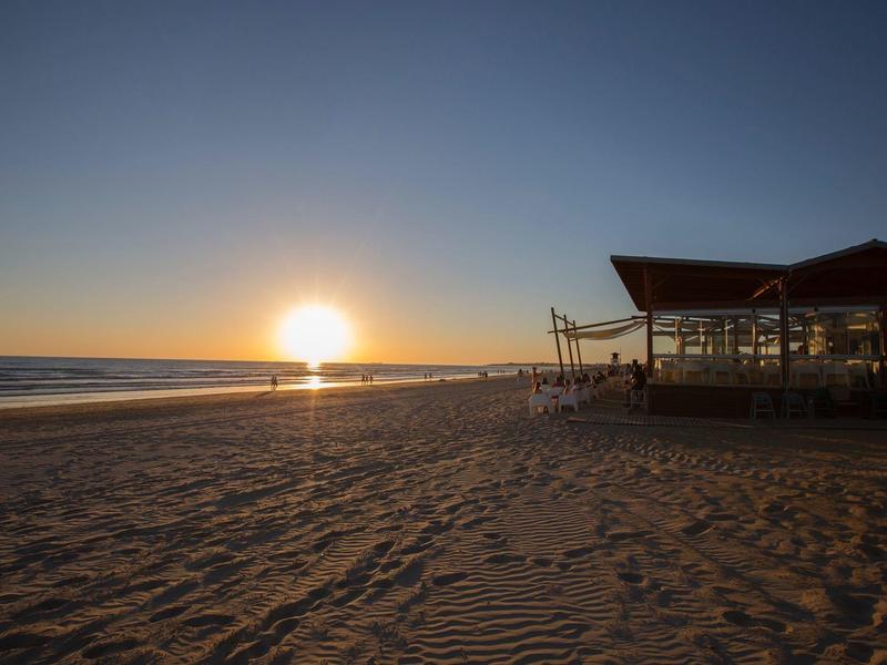 Sonnenuntergang am Strand mit weichgezeichnetem Sand und einem schattigen Pavillon rechts.