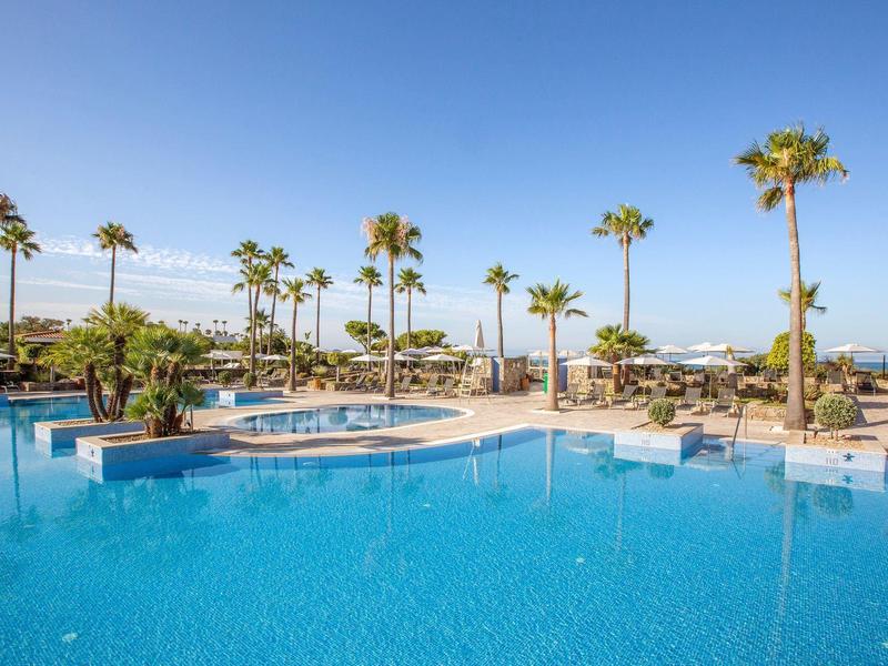 Großer blauer Pool mit Palmen, Sonnenliegen und klarem Himmel im Hintergrund.