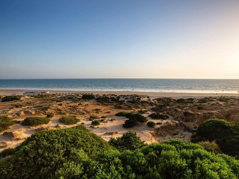 Küstenlandschaft mit Sand, Felsen, grünem Buschwerk und ruhigem, blauem Meer unter klarem Himmel.