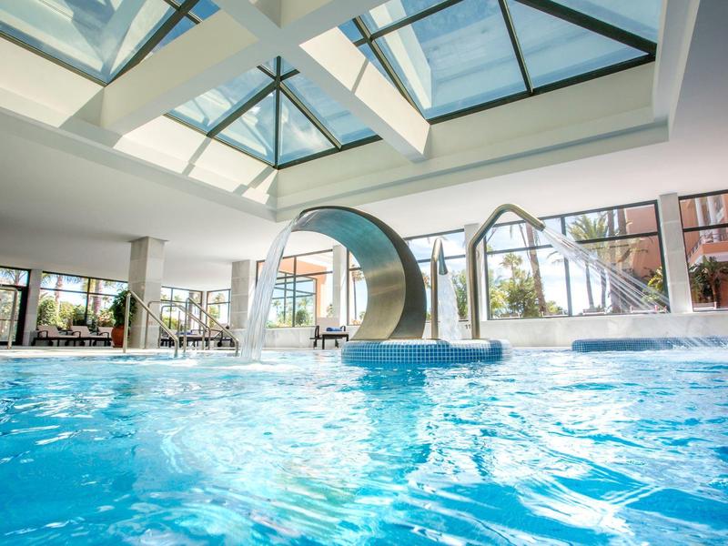 Helles Hallenbad mit großem Fenster, Wasserrutsche und klarem blauem Wasser unter Glasdach.