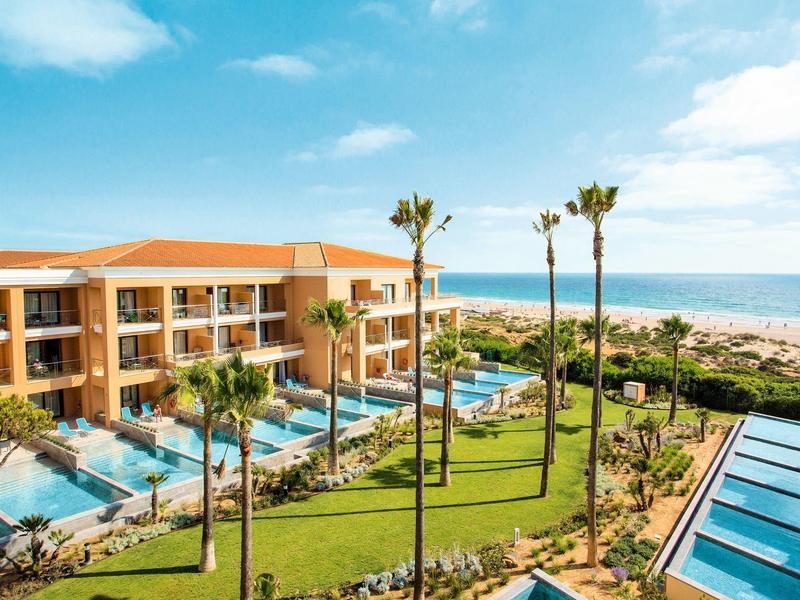 Hotelanlage mit Pool, Palmen und Blick auf den Strand und das blaue Meer unter leicht bewölktem Himmel.