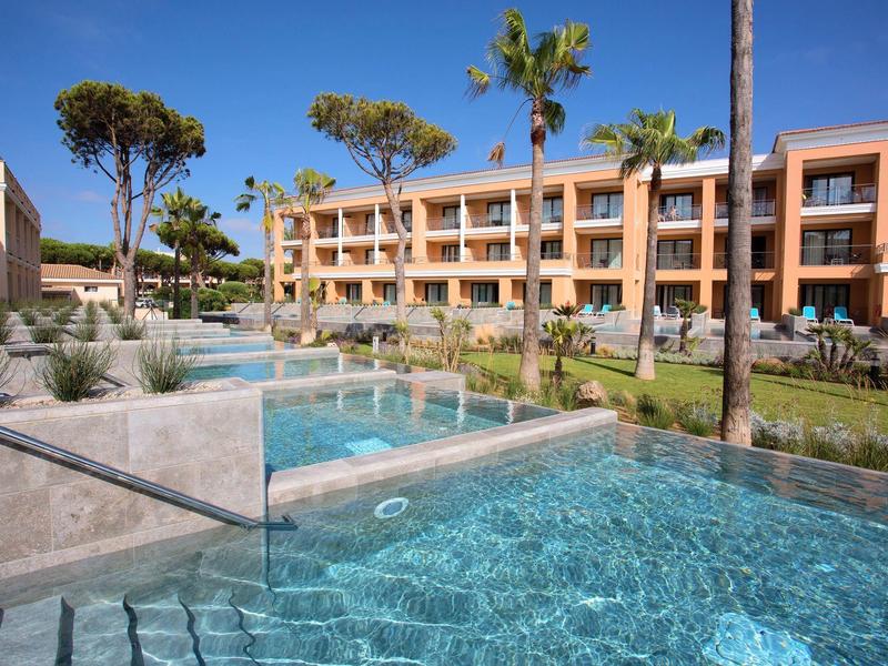 Moderner Hotelkomplex mit mehrstöckigen Gebäuden, klarem Pool und Palmen im sonnigen Ambiente.