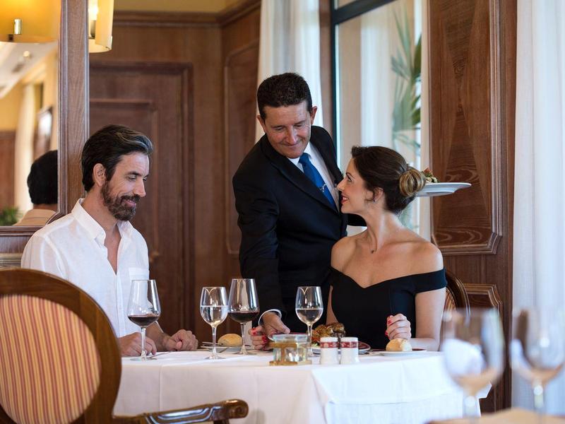Drei Personen am Tisch in elegantem Restaurant, zwei sitzen, einer serviert speisen und Getränke.