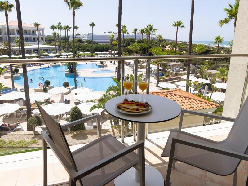Balkon mit Tisch und zwei Stühlen, Blick auf Pool, Palmen und weiße Sonnenschirme bei klarem Himmel.