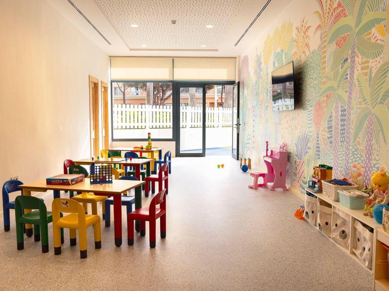 Heller Kinderspielraum mit bunten Stühlen, Tischen, Spielzeug und großer Wandillustration.