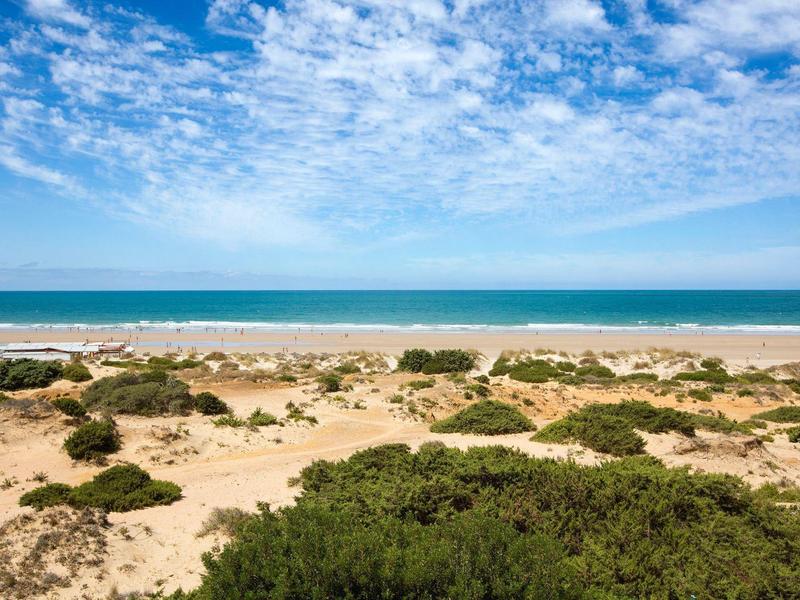 Strand mit Sanddünen, grünen Büschen und blauem Meer unter teils bewölktem Himmel.