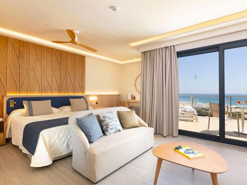 Helles Hotelzimmer mit Holzwand, Doppelbett, Couch, großem Fenster und Meerblick.