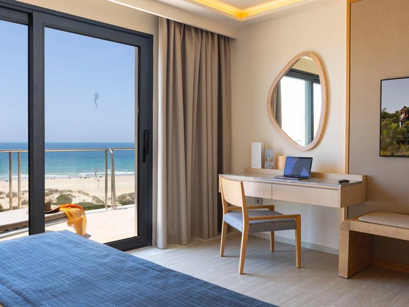 Helles Zimmer mit Schreibtisch, Stuhl, rundem Spiegel, Bett und Meerblick durch große Fenster.