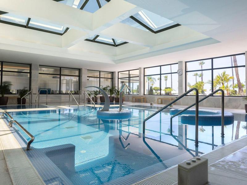 Hell beleuchtetes Hallenbad mit großem rechteckigem Pool, Pflanzen und Glasfenstern rundherum.