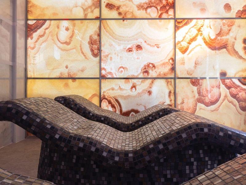 Zwei wellenförmige, gepolsterte Liegen vor einer Wand mit großformatigen, bunten Marmorplatten.