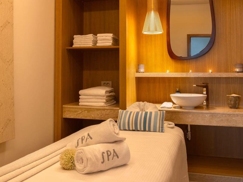 Gemütlicher Spa-Raum mit Massageliege, weißen Handtüchern, Holzregalen und einem Waschbecken mit Spiegel.