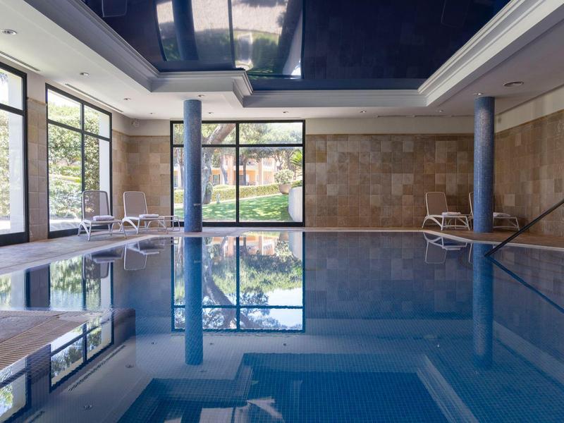 Helles Hallenbad mit großen Fenstern, blauen Säulen und Liegestühlen am Beckenrand.