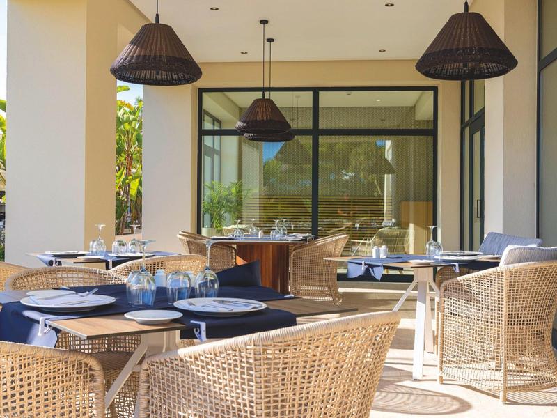 Offener Gastraum mit Korbstühlen, Holz- und Metalltischen, Lampen und großen Fenstern, viel Tageslicht.