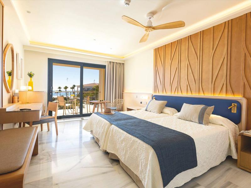 Helles Hotelzimmer mit großem Bett, blauem Überwurf, Holzverkleidung und Balkon mit Meerblick.