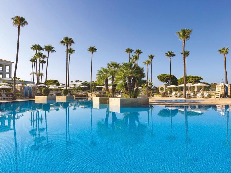 Großer blauer Pool mit Palmen und Liegestühlen bei klarem Himmel in einer Ferienanlage.