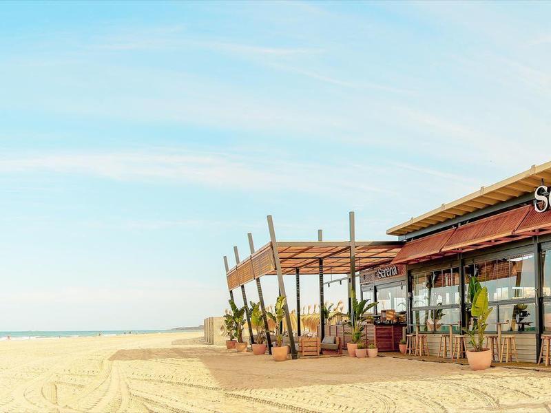 Strandbar mit Holzterrasse am sandigen Strand unter klarem, blauem Himmel.