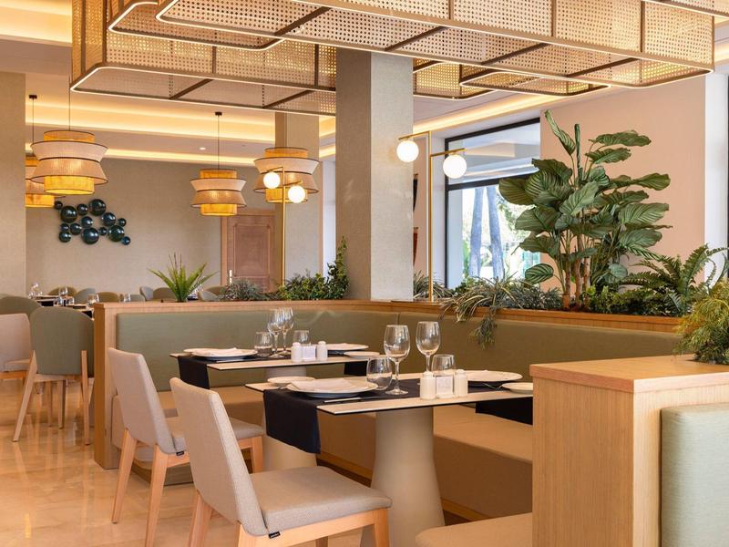 Helles Restaurant mit beigen Stühlen, Holzmöbeln, Pflanzen und moderner Deckenbeleuchtung.